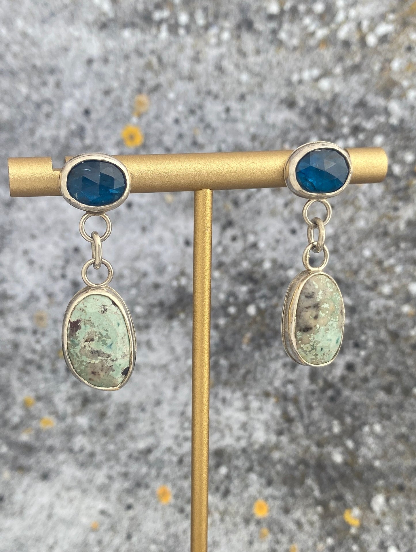 Pair of earrings with blue and green stones on a gold stand against a textured gray background
