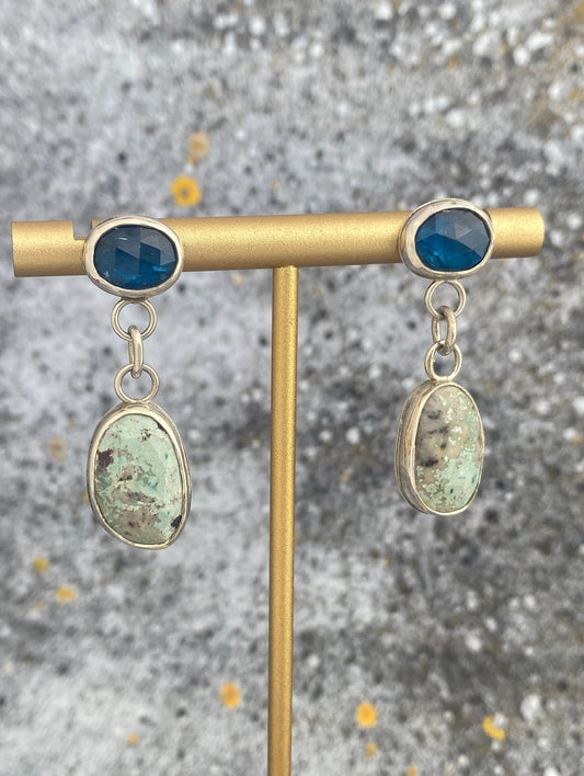 Pair of earrings with blue and green stones on a gold stand against a textured gray background