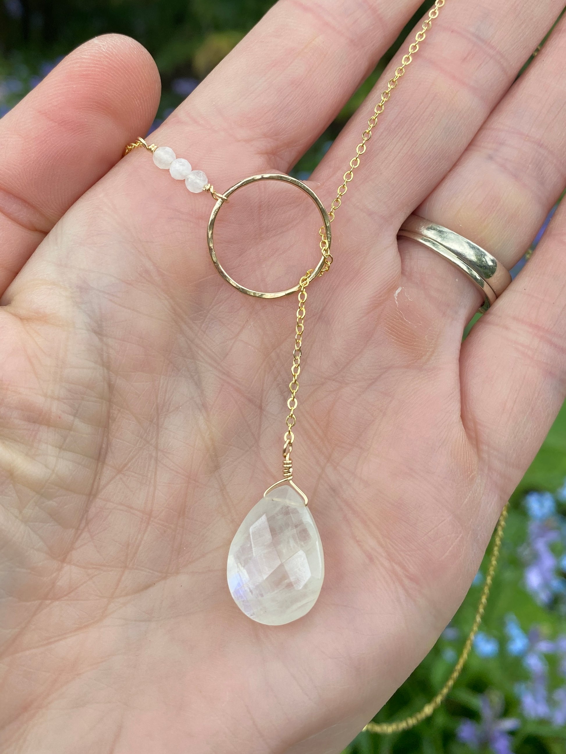 Gold lariat necklace with a teardrop-shaped moonstone pendant held in a hand against a blurred natural background