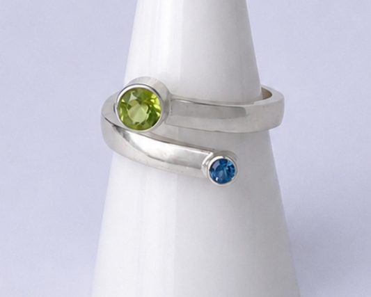 Silver ring with green and blue gemstones on a white cone-shaped stand against a light purple background